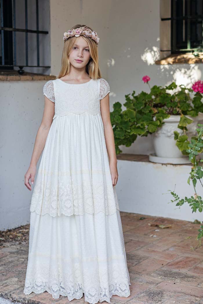 Niña con un vestido blanco de encaje de pie en un patio de piedra cerca de macetas con flores y vegetación.