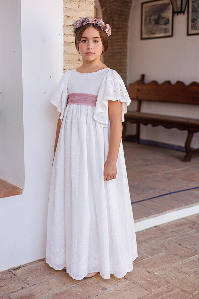 Niña con un vestido blanco y una faja rosa de pie junto a una pared blanca en un patio.