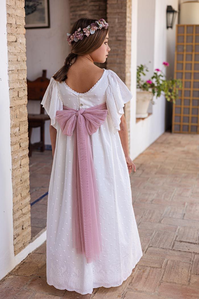 Una niña con un vestido blanco, una faja rosa y una diadema floral, de pie en un porche y mirando hacia otro lado.