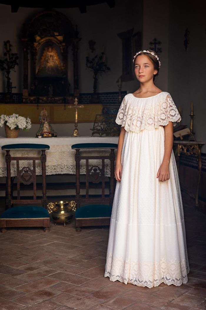 Mujer con un vestido de encaje blanco de pie en una habitación con poca luz, con mesas y decoración religiosa.