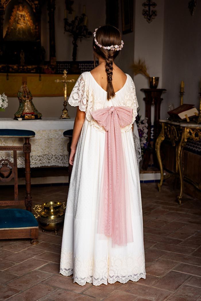 Niña con un vestido blanco de encaje y una faja rosa, vista de espaldas en una habitación con un altar.
