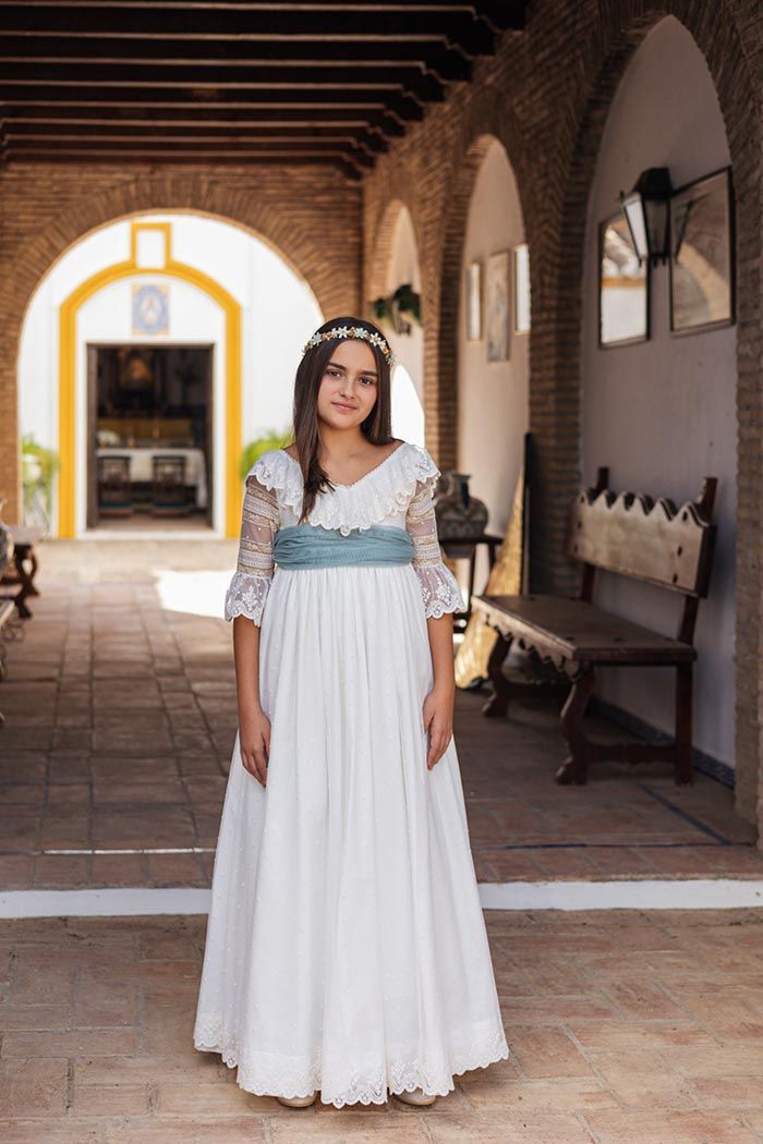 Mujer con vestido blanco de pie en un pasillo de piedra iluminado por el sol, con arcos y bancos.