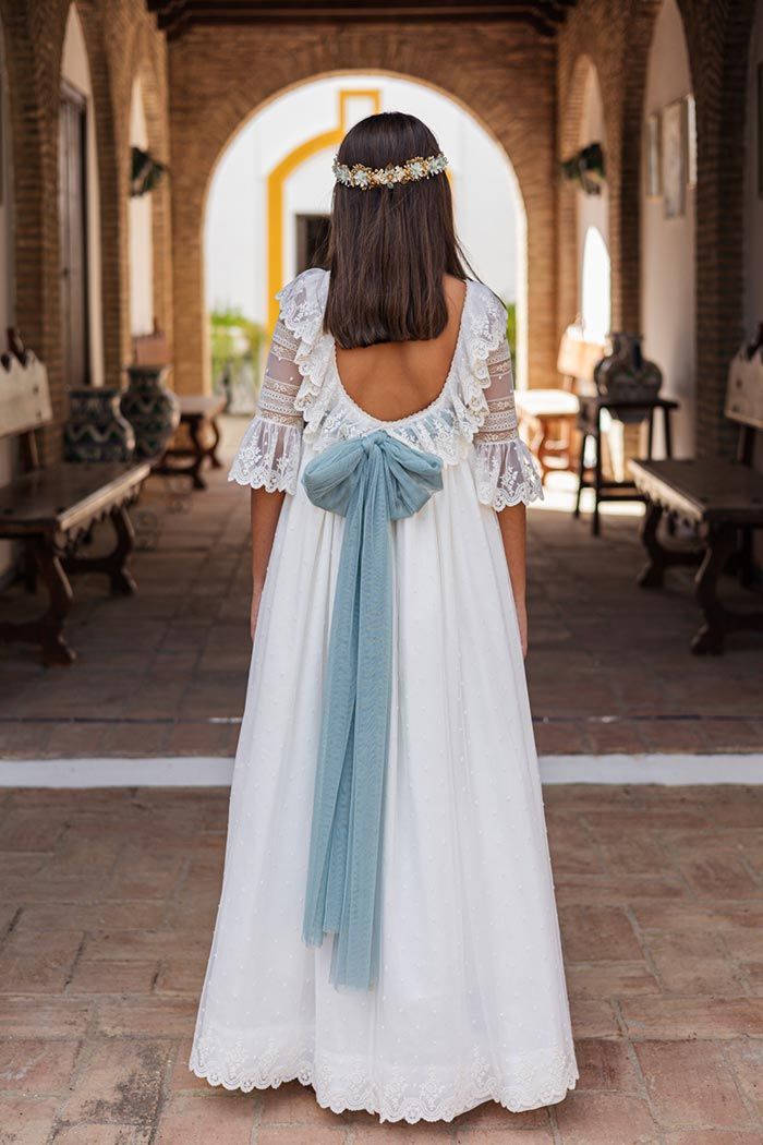 Mujer con un vestido blanco y un largo lazo azul, de pie en el pasillo de una iglesia, de espaldas a la cámara.