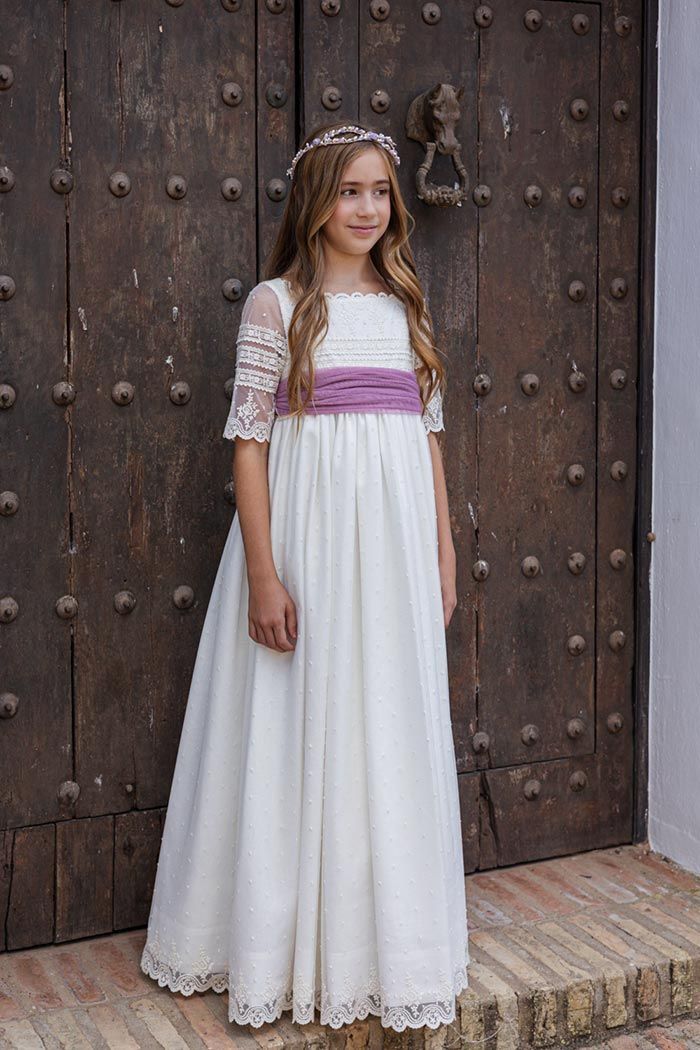 Una persona vestida de blanco con una faja morada está de pie frente a una gran puerta de madera.