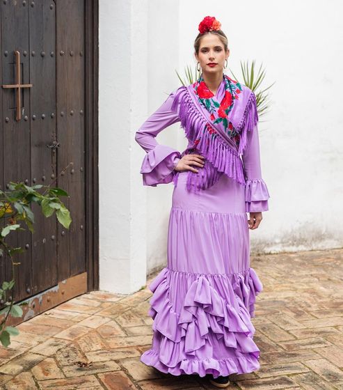 Mujer con un vestido lila con volantes de pie al aire libre junto a una pared blanca y una puerta de madera.