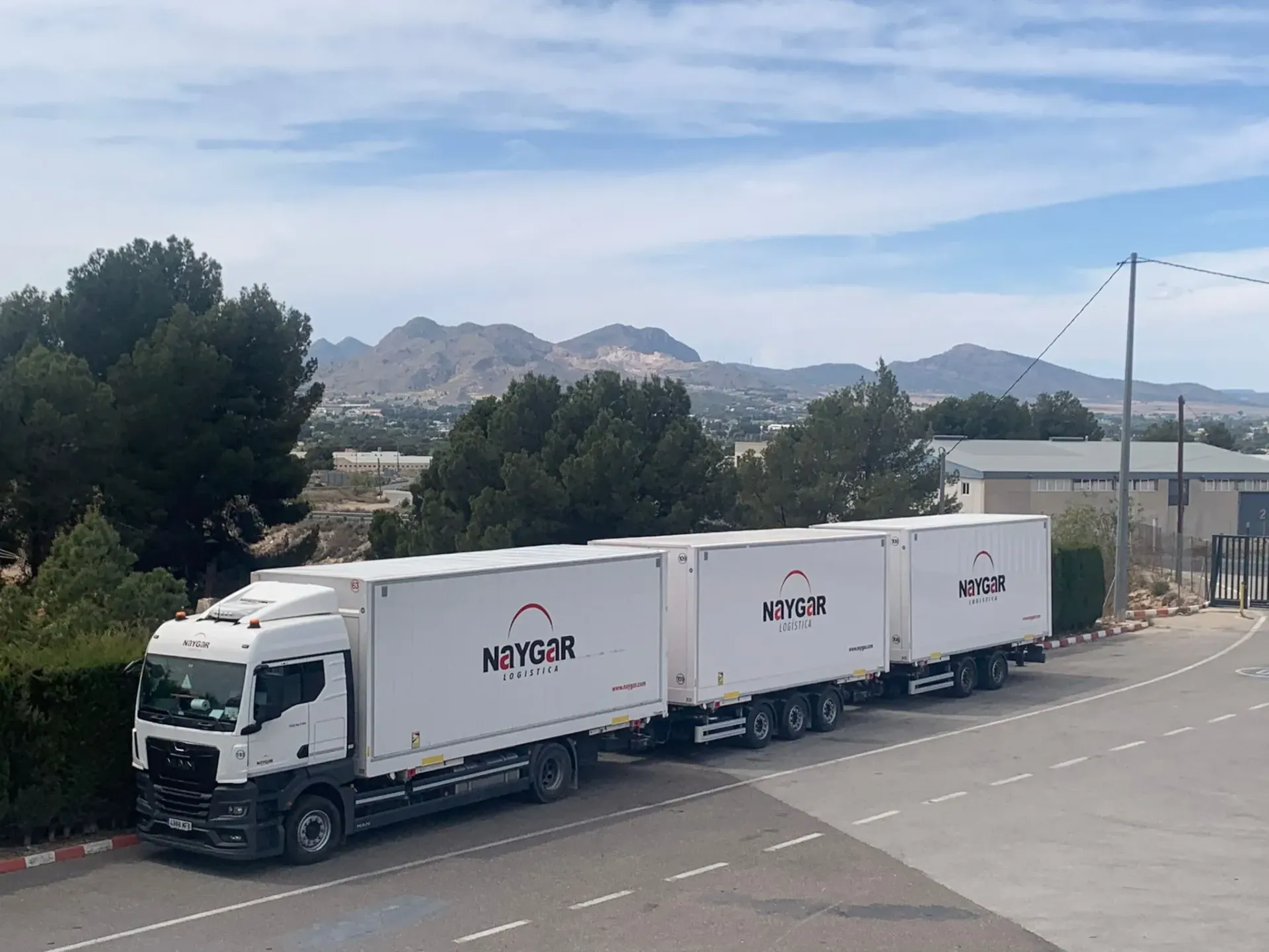 Camión semirremolque blanco con tres remolques conectados estacionado en una carretera, montañas al fondo.