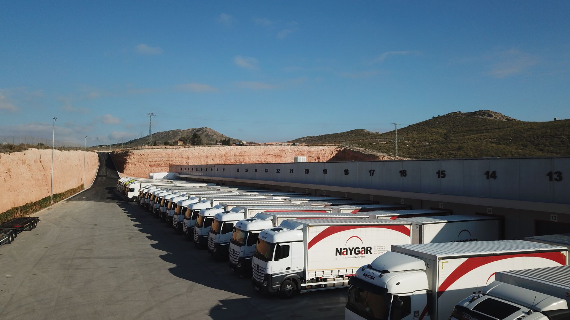 Camiones alineados en un muelle de carga bajo un cielo azul, algunos con el logotipo