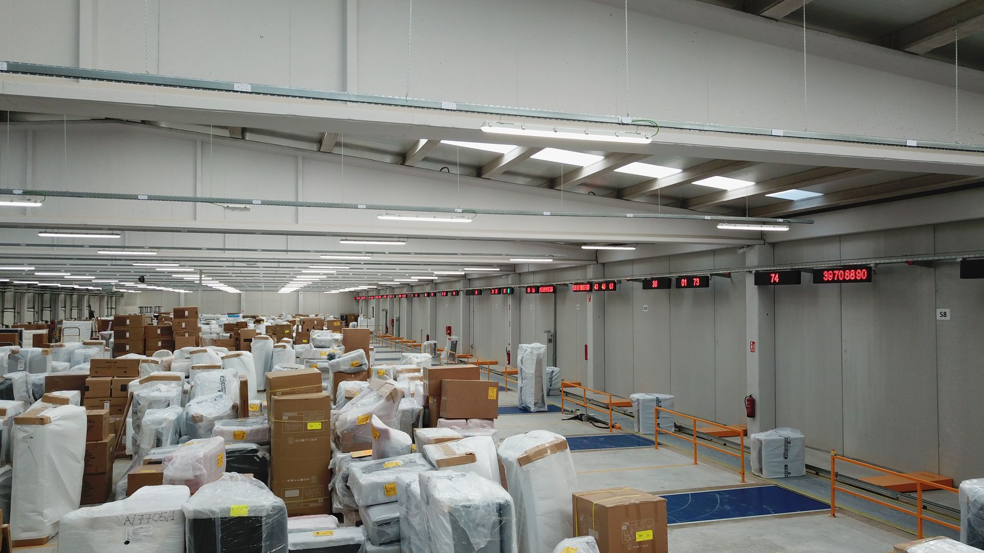 Interior de almacén con cajas apiladas e iluminación superior, pantallas digitales y una cinta transportadora.