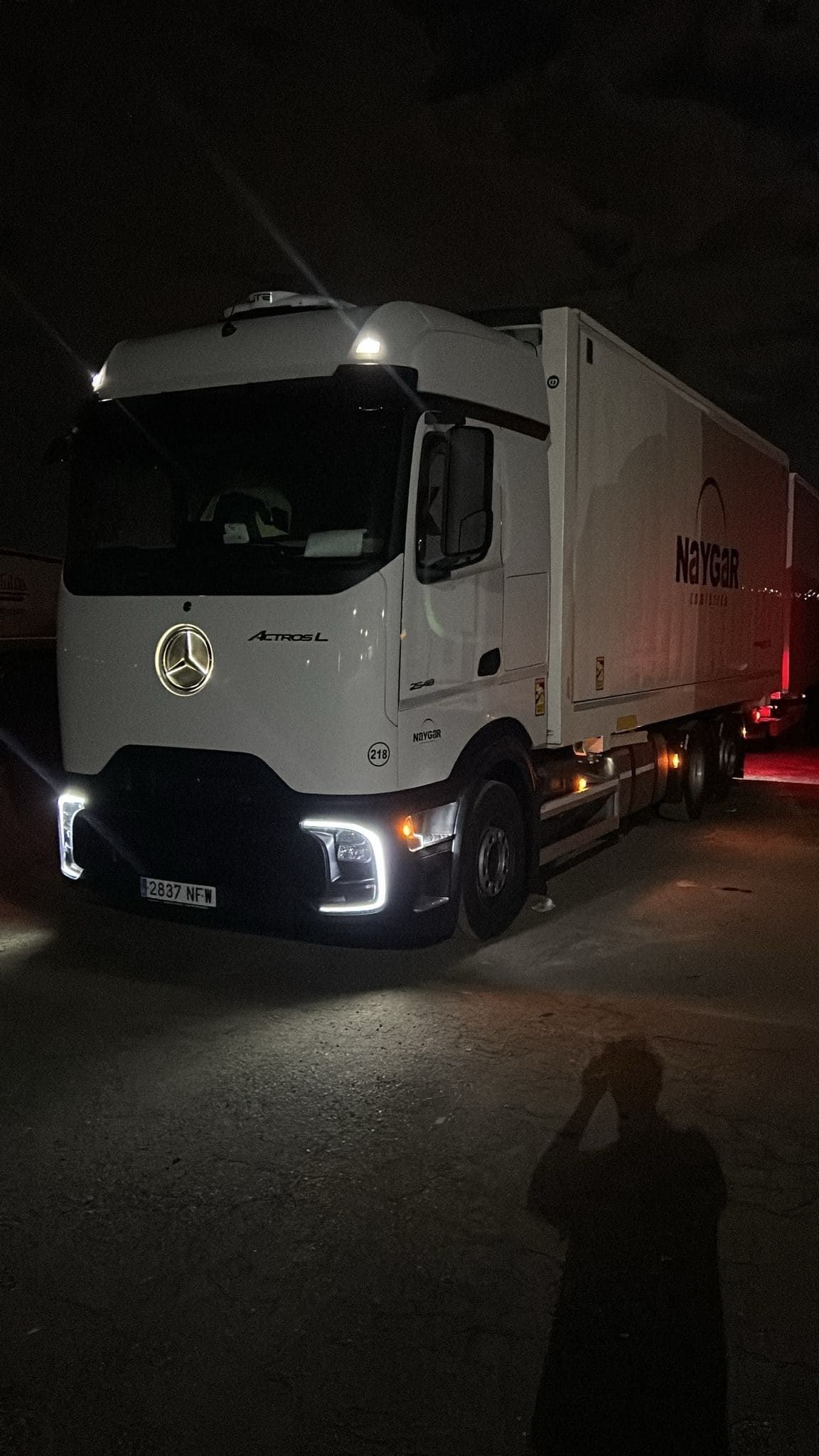Camión semirremolque blanco estacionado de noche, con los faros y las luces de la cabina encendidos. Una persona toma una foto.