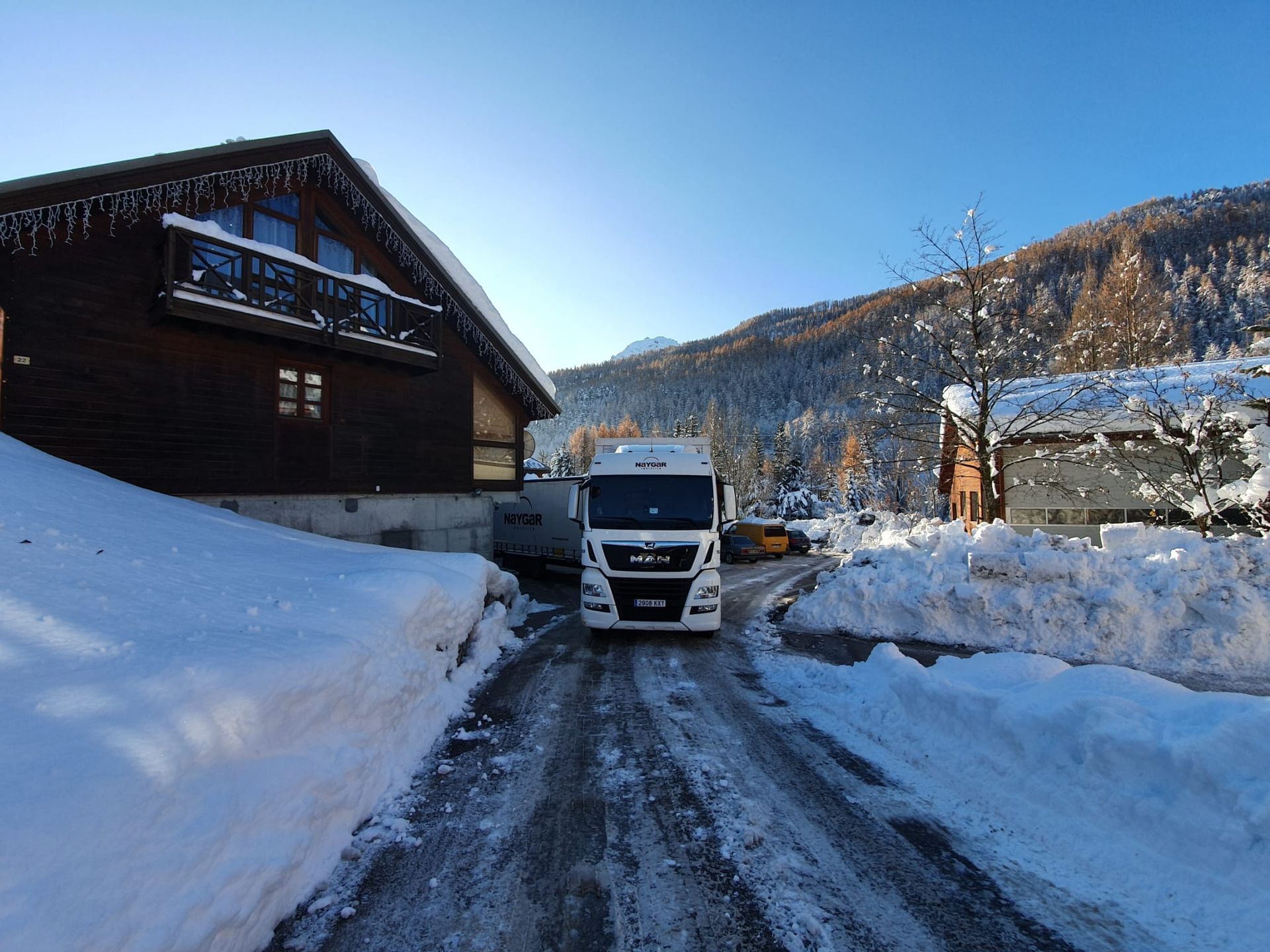 Camión circulando por una carretera nevada en un pueblo de montaña, entre edificios de madera con nieve.