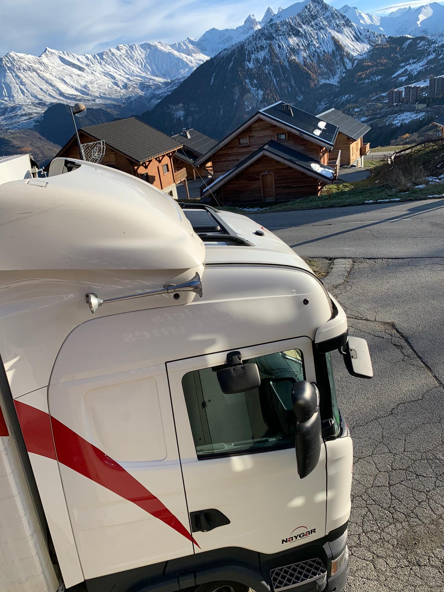 Camión blanco con franja roja estacionado frente a montañas nevadas y chalets de madera.
