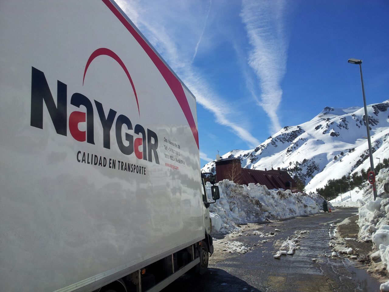 Camión Naygar blanco en entorno de montaña nevada, logotipo visible, día soleado.