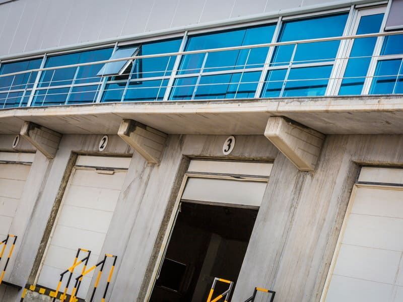 Muelles de carga con puertas parcialmente abiertas, bajo ventanas tintadas de azul. Estructura de hormigón.