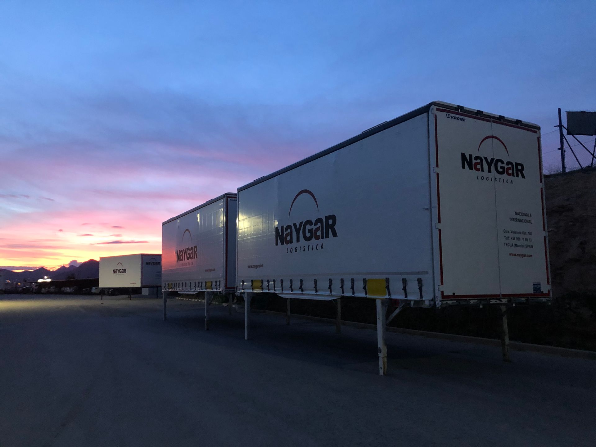 Remolques semirremolque Naygar blancos estacionados en un estacionamiento, con el cielo del atardecer de fondo.