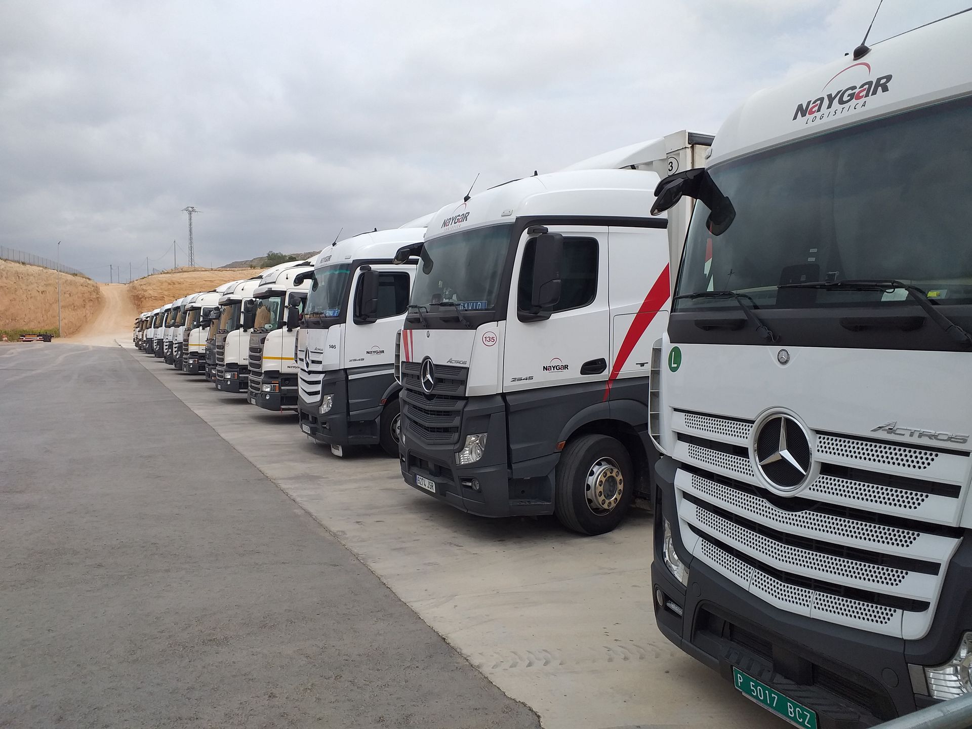 Una fila de camiones semirremolques blancos con detalles en rojo estacionados en un lote de concreto, bajo un cielo nublado.