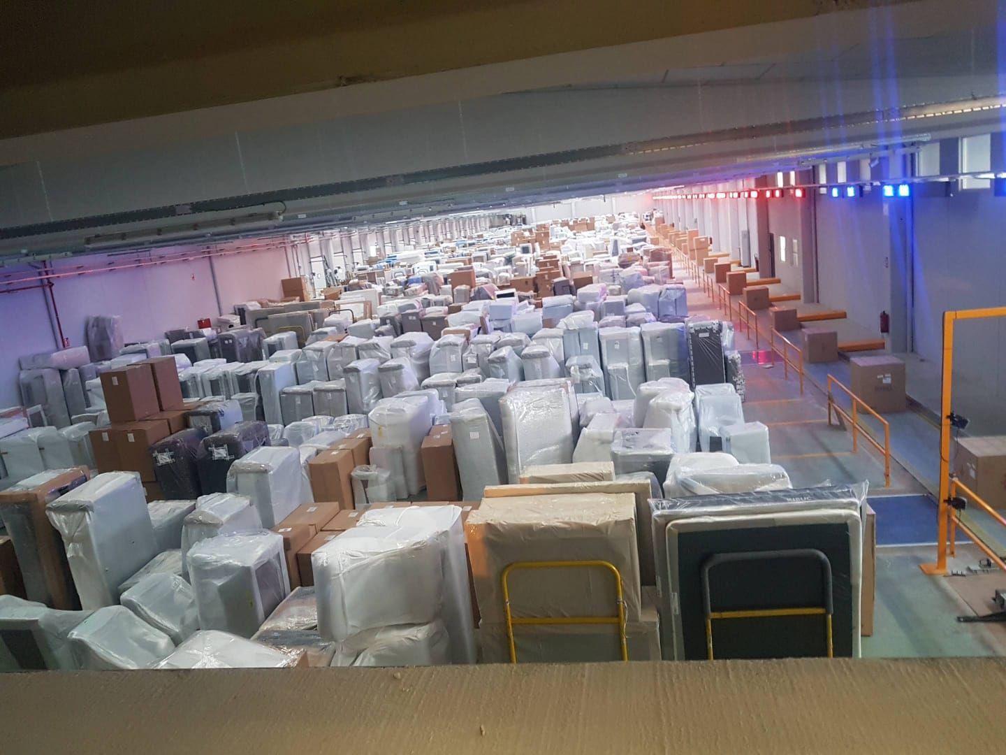 Almacén lleno de cajas y paquetes envueltos; vista interior con iluminación cenital.