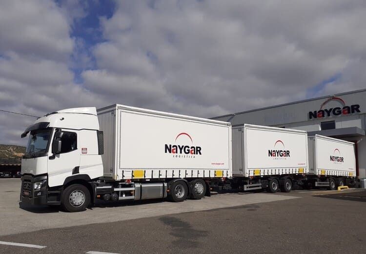 Camión de transporte Naygar de color blanco con tres remolques conectados, estacionado frente a un edificio.