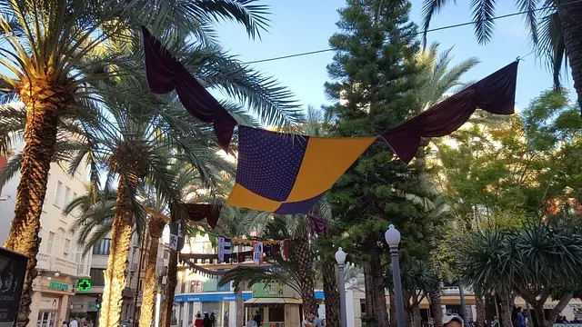 Una bandera morada y amarilla cuelga entre dos palmeras en un parque.