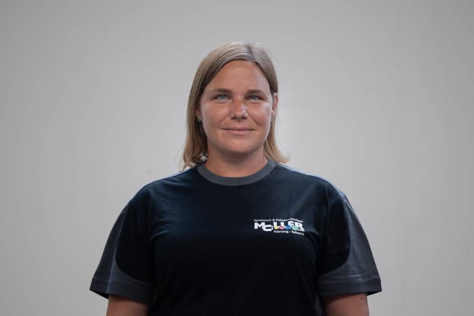 Frau in einem dunklen T-Shirt mit Logo vor hellgrauem Hintergrund; lächelnd, blickt in die Kamera.