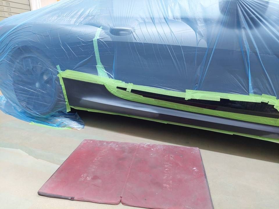 Auto ist mit blauer Folie und grünem Klebeband für die Lackierung abgeklebt; davor liegt ein roter Schleifklotz bereit.