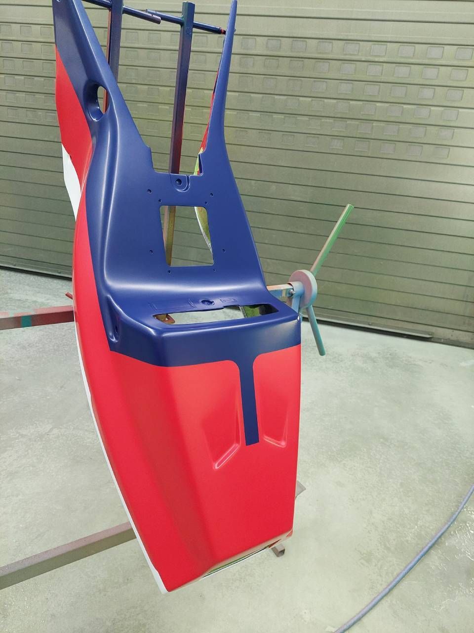 Ein rot, weiss und blau lackierter Bootsrumpf hängt in einer Spritzkabine an einem Gestell.