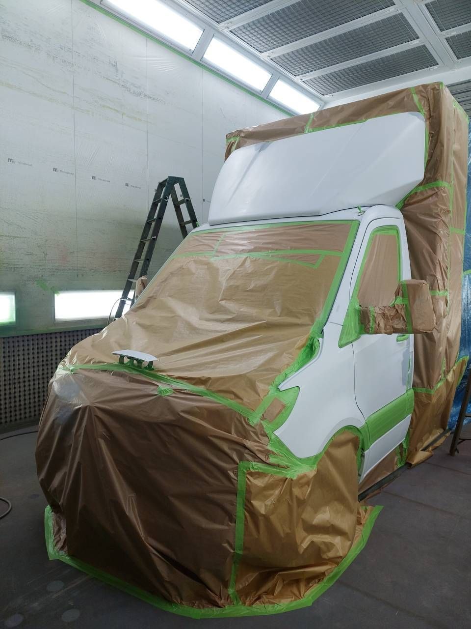 Ein weisser Lieferwagen in einer Lackierkabine, teilweise mit braunem Abdeckpapier und grünem Klebeband beklebt.