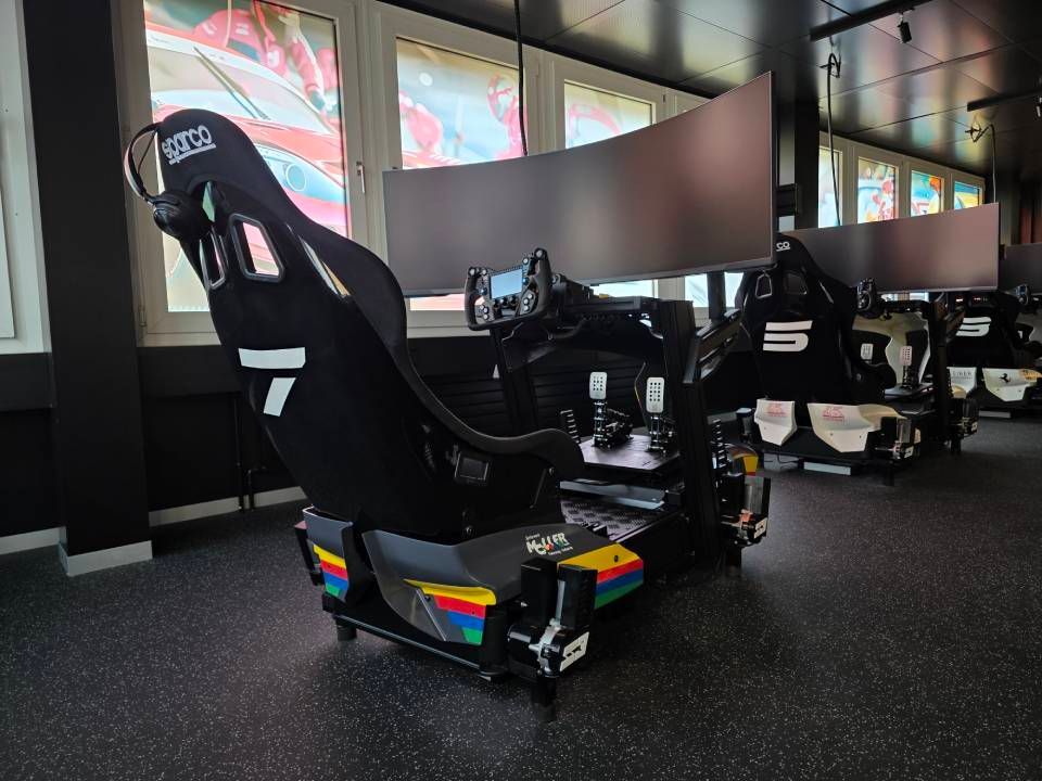 Rennsimulator mit schwarzem Sitz und gebogenem Monitor, in einem Raum mit anderen Simulatoren und Fenstern.