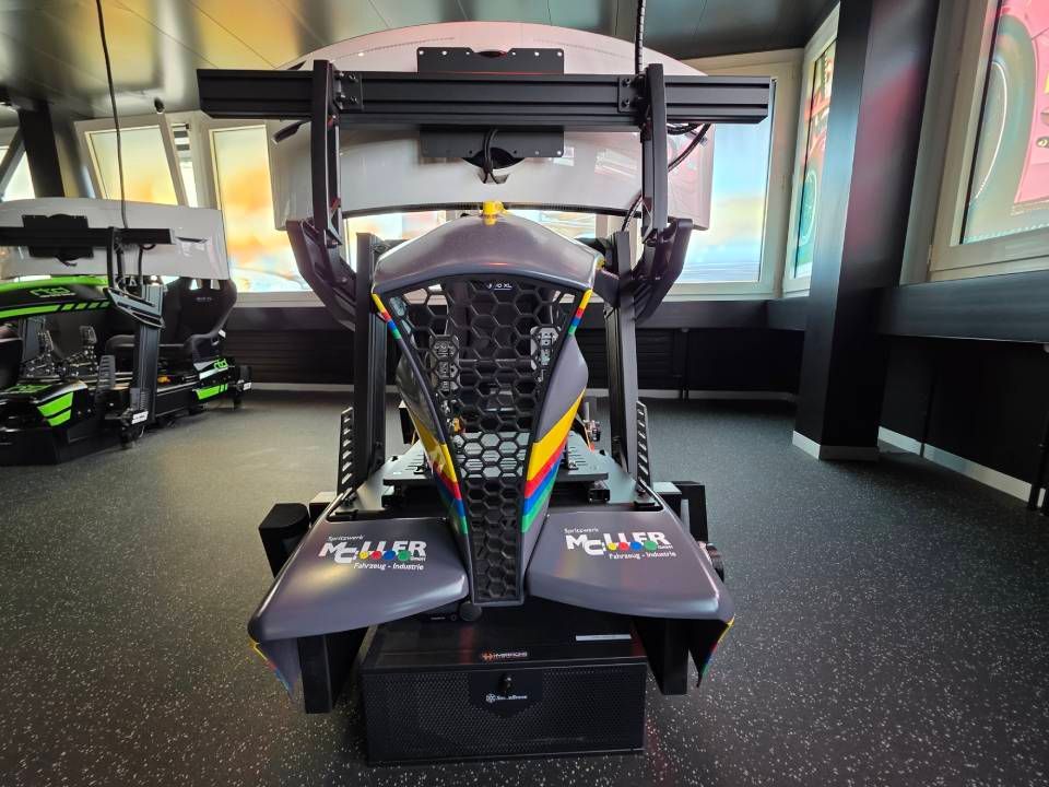 Rennsimulator mit grauen, schwarzen und regenbogenfarbenen Akzenten in einem Raum mit grossem Fenster und anderen Simulatoren.