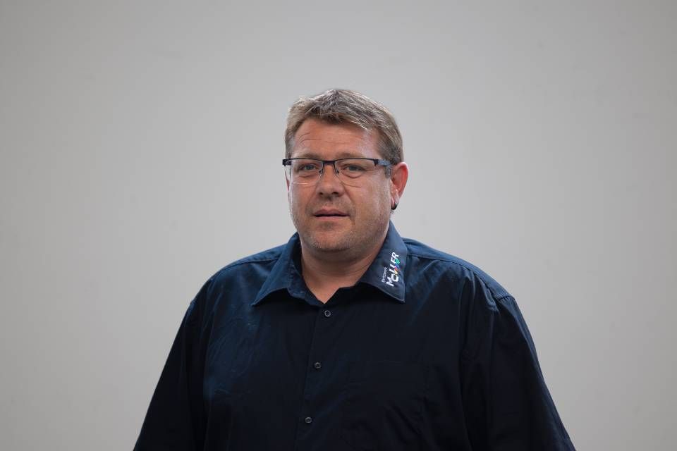 Mann mit Brille und dunklem Hemd, Blick nach vorn, vor hellgrauem Hintergrund.