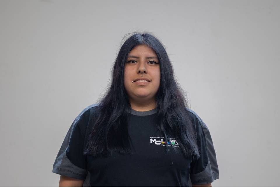 Eine Frau mit langen dunklen Haaren, die ein dunkelblaues T-Shirt trägt, steht lächelnd vor einer weissen Wand.