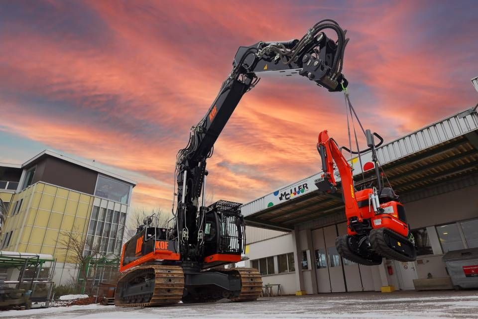 Großer orangefarbener Bagger hebt einen kleineren Bagger vor einem dramatischen Sonnenuntergangshimmel auf Baustelle.