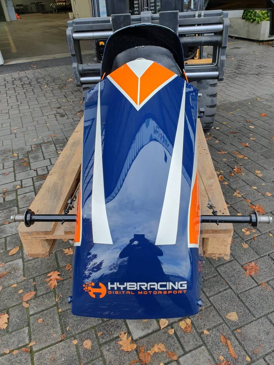 Blauer und orangefarbener Rennwagen auf einer Palette, Logo „HYBRACING DIGITAL MOTORSPORT“ sichtbar.