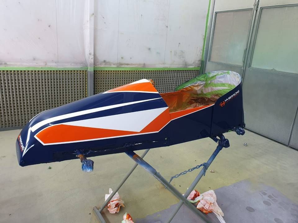 Eine blau, orange und weiss lackierte Fahrzeugkarosserie steht auf einem Ständer in einer Werkstatt.