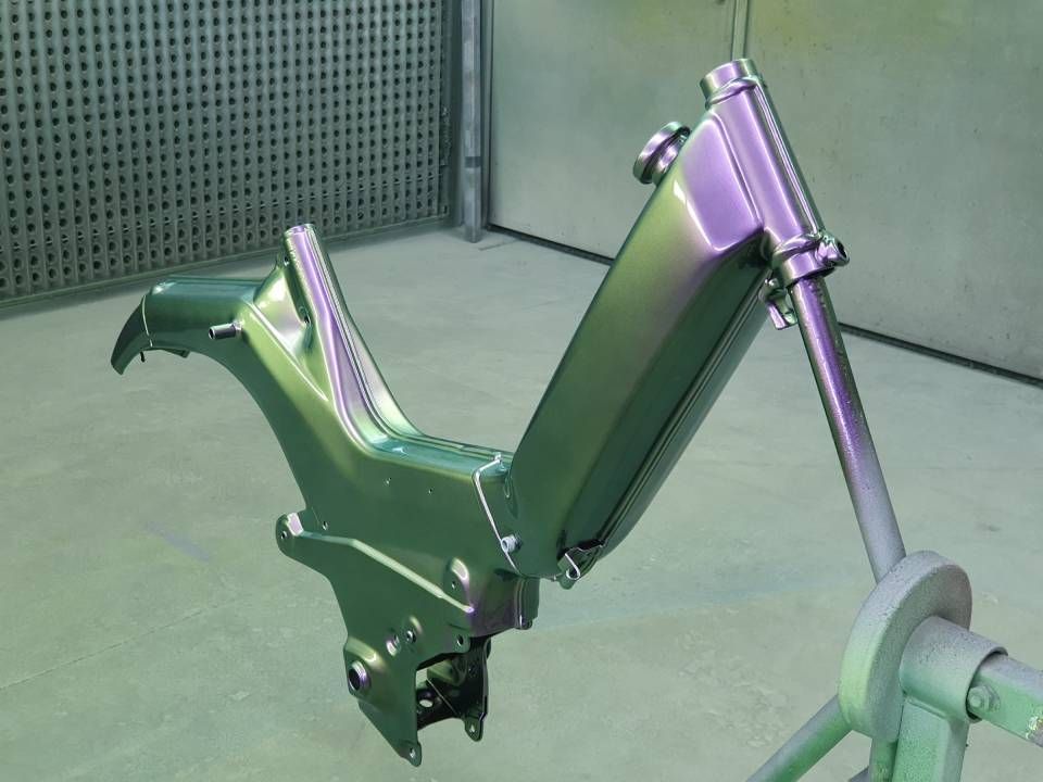 Ein grün-violett schimmernder Motorradrahmen wird in einer Lackierkabine lackiert.