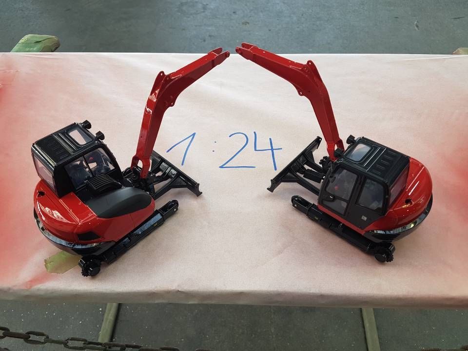 Zwei rot-schwarze Spielzeugbagger stehen sich mit erhobenen Armen auf rosa Fläche gegenüber; dazwischen die Zahl „1:24“.