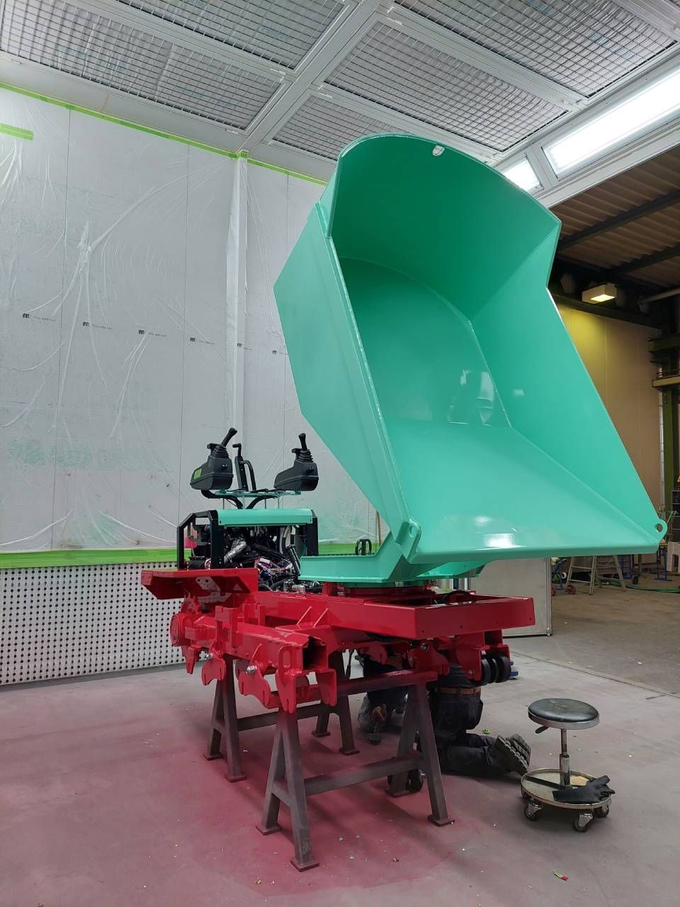 Grüne Muldenkipperschaufel angehoben, rotes Fahrgestell, dunkler Motor, in einer Werkstatt.