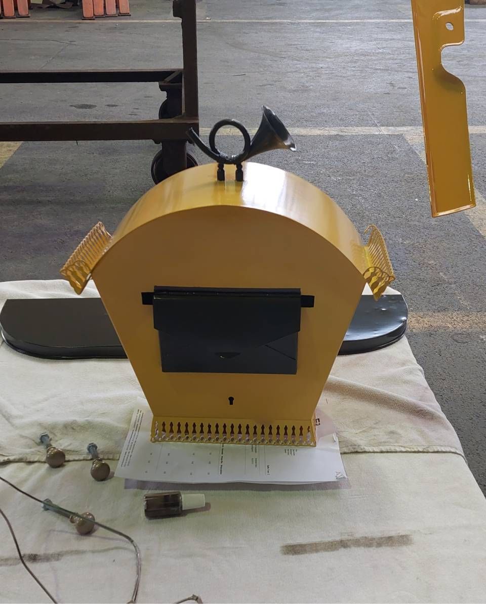 Ein gelber Metallbriefkasten mit Hornverzierung, schwarzem Schlitz und detailliertem Sockel steht auf einem Tisch.