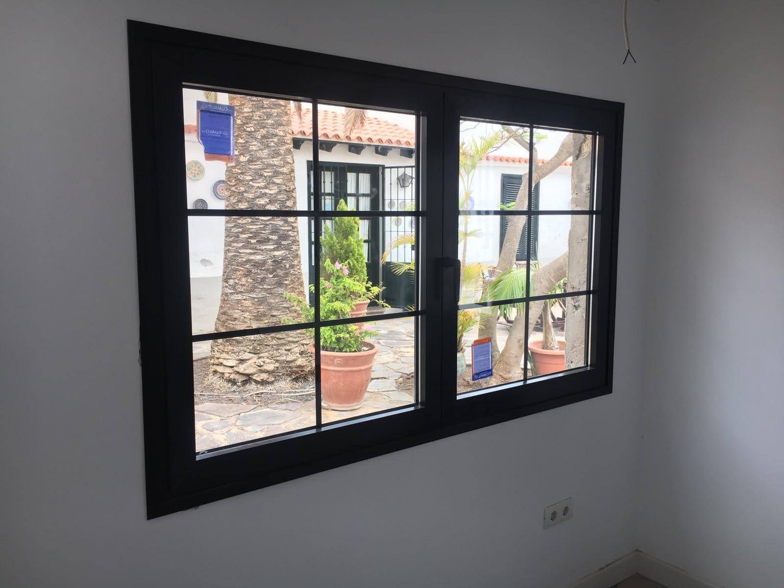 Ventana con marco negro y paneles de rejilla, con vista a un patio con plantas, árboles y edificios blancos.