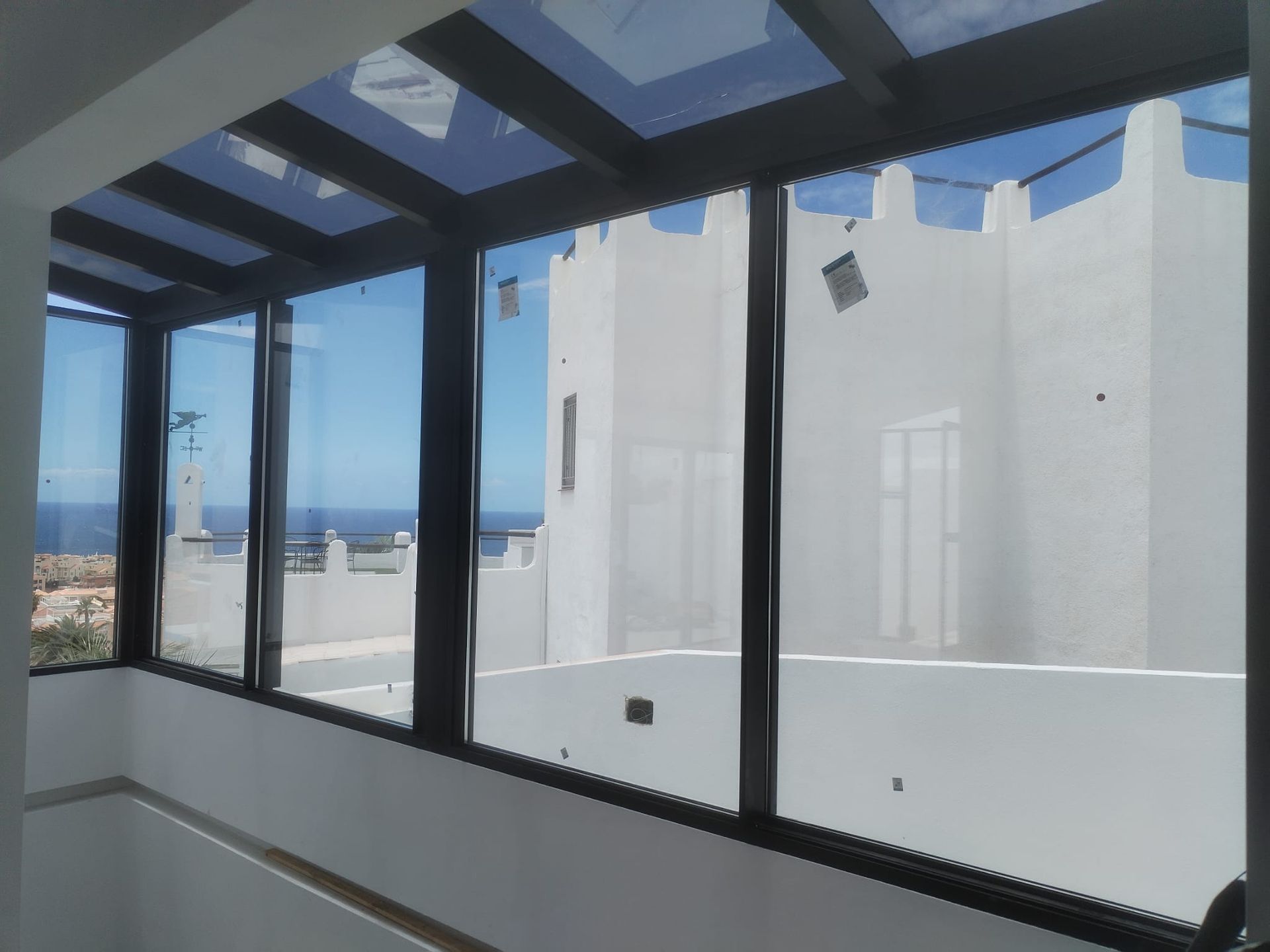 Balcón acristalado con marcos negros y vista al océano y al edificio blanco bajo un cielo azul brillante.