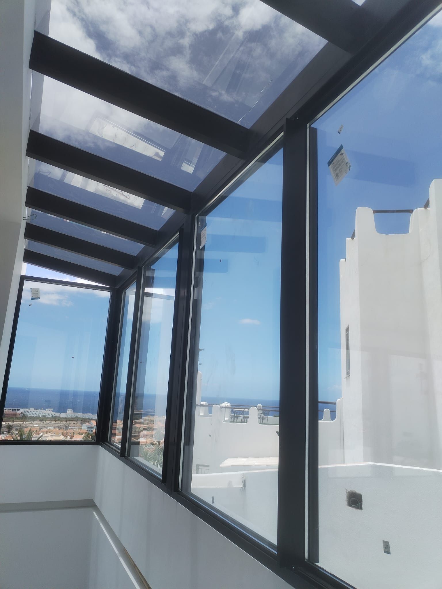 Pasarela con paredes de vidrio y techo parcialmente de vidrio, con vista a una ciudad, al mar y a un edificio blanco en un día soleado.