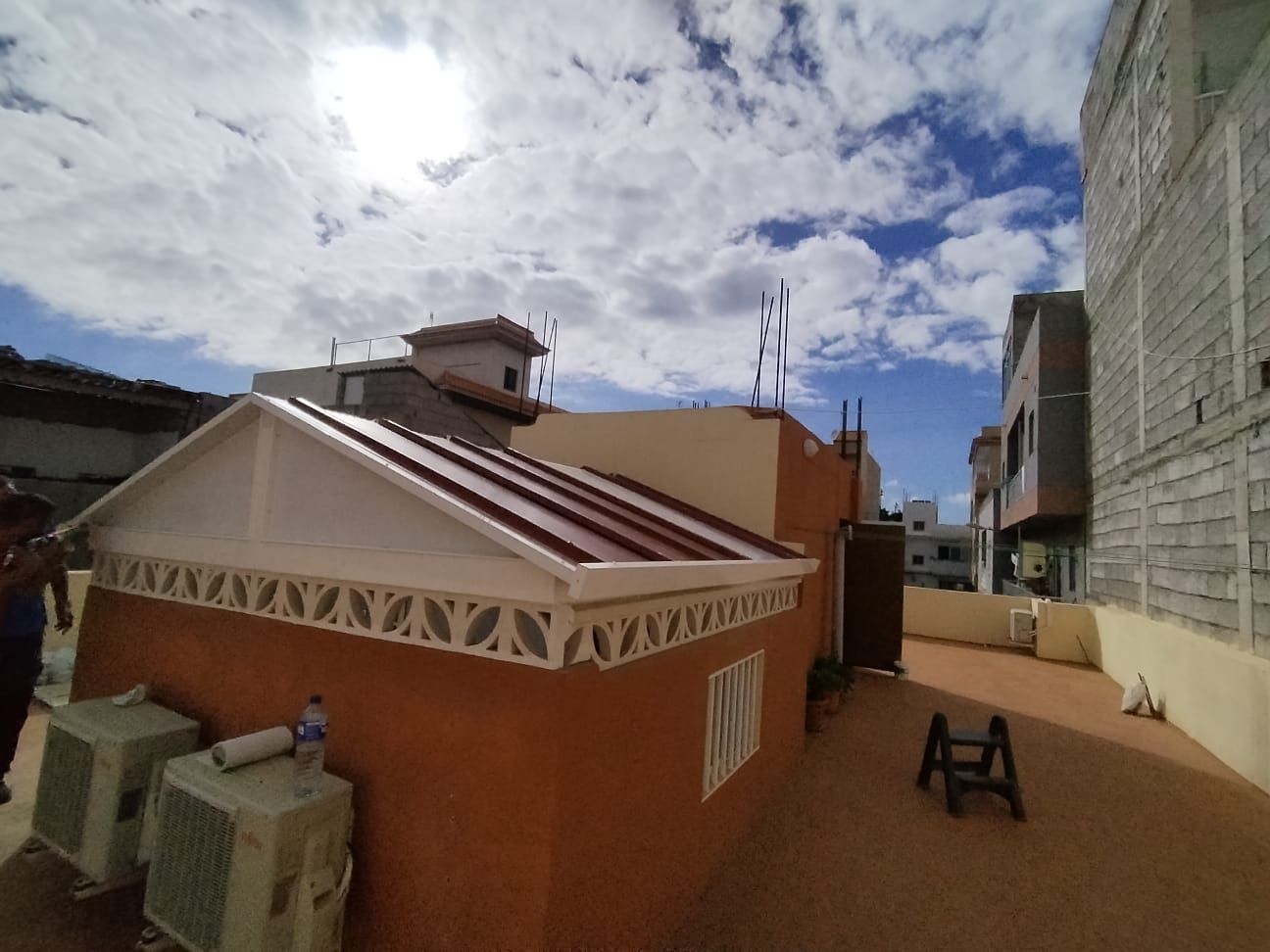 Vista de los tejados con un pequeño edificio con un techo rojo en ángulo, unidades de aire acondicionado y un cielo parcialmente nublado.
