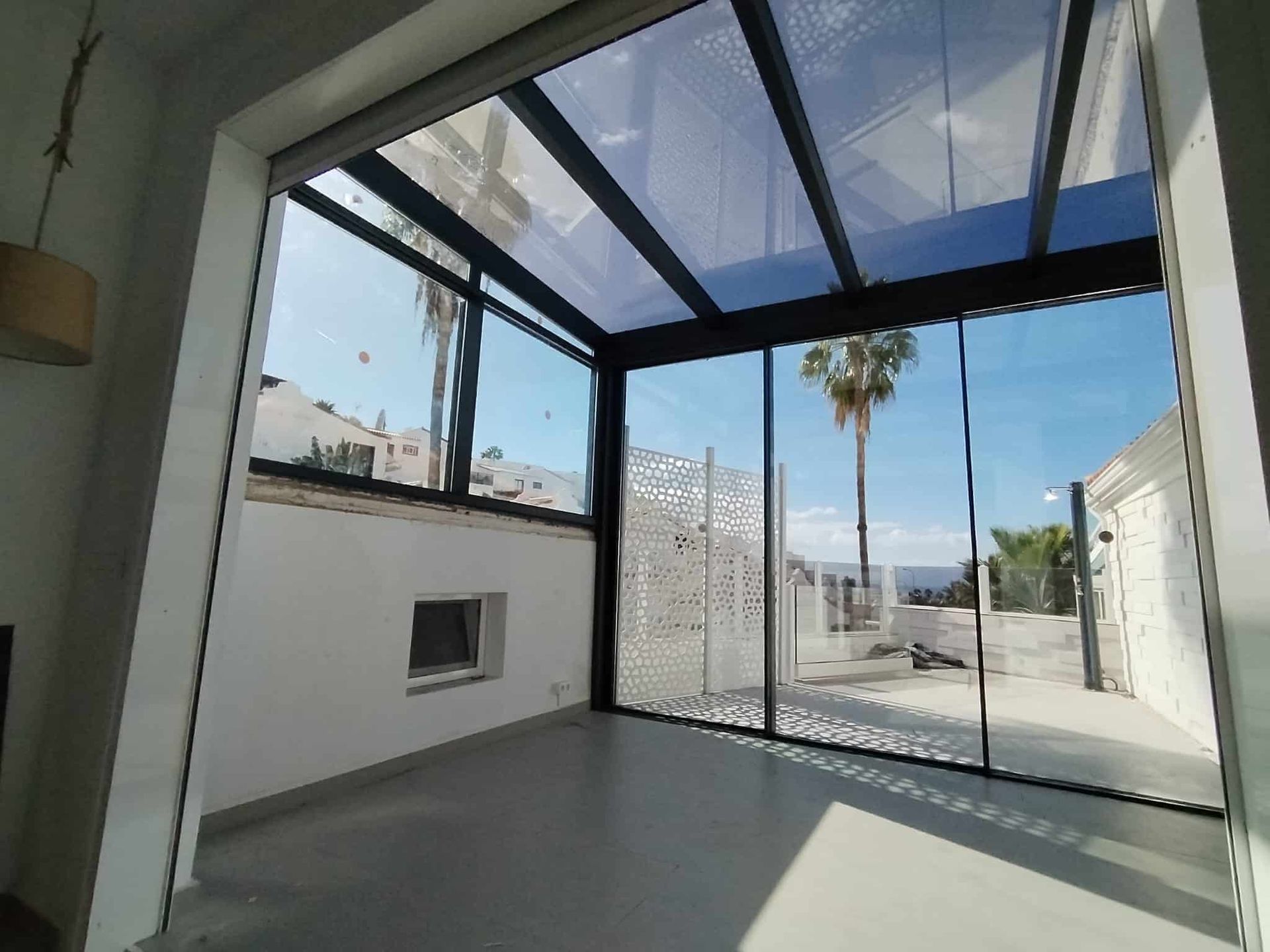 Terraza acristalada con techo transparente. Palmeras y cielo azul visibles en el exterior.