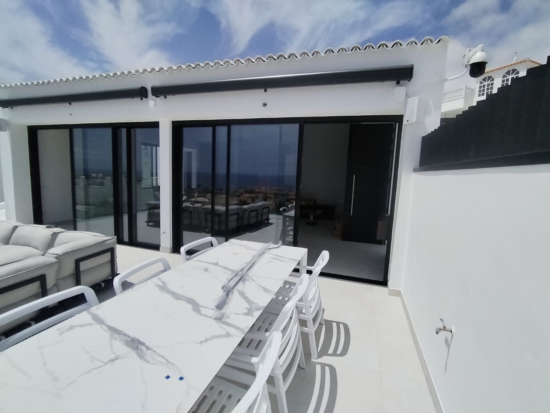 Patio con mesa y sillas blancas, puertas corredizas de vidrio al interior, vista al mar.