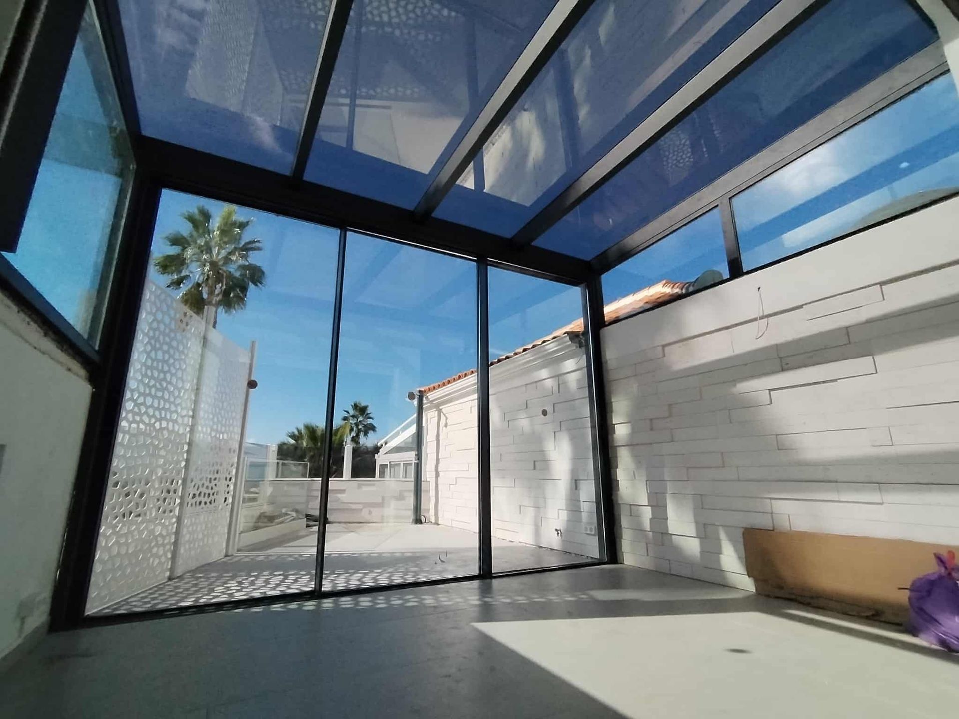 Terraza acristalada con paredes y techo de cristal, con vistas a un patio y al cielo azul.