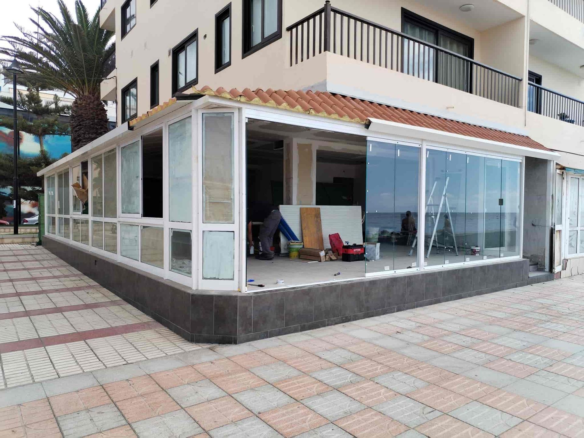 Estructura acristalada adosada a un edificio, posiblemente un restaurante. Trabajadores en el interior; entorno costero.