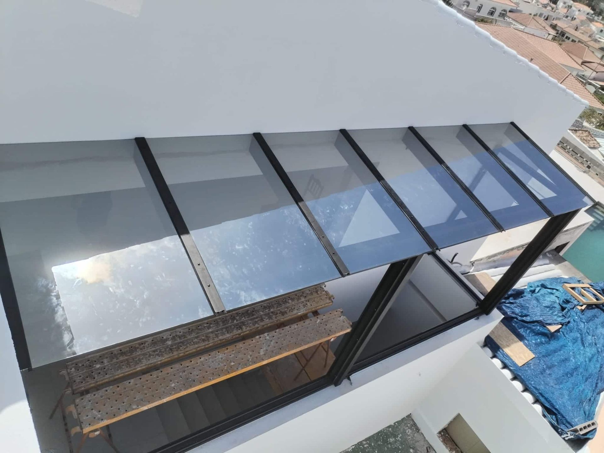 Toldo de cristal en un edificio blanco, sobre un balcón. Marco negro, paneles transparentes, luz solar intensa, vista exterior.