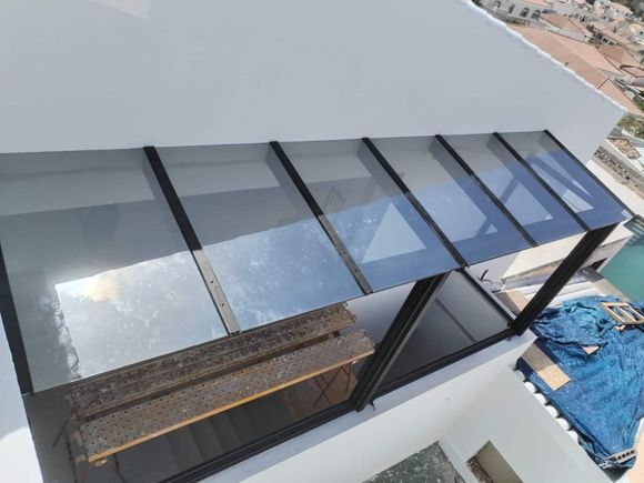 Toldo de cristal en un edificio blanco, sobre un balcón. Marco negro, paneles transparentes, luz solar intensa, vista exterior.