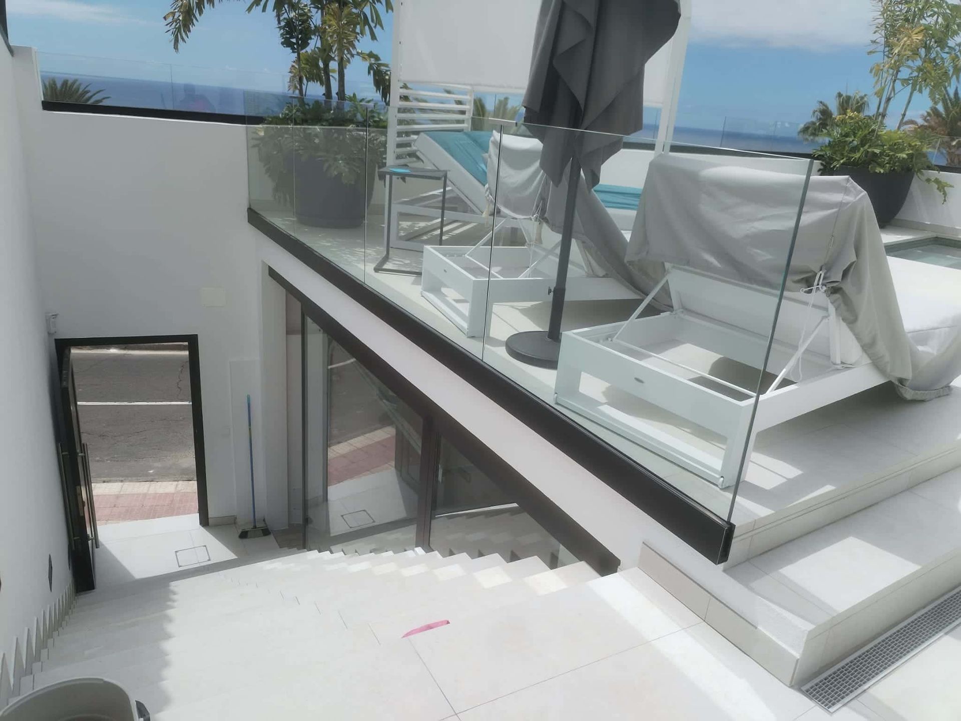 Patio en la azotea con muebles blancos, barandilla de cristal, piscina y escaleras. Día soleado y luminoso con cielo azul.