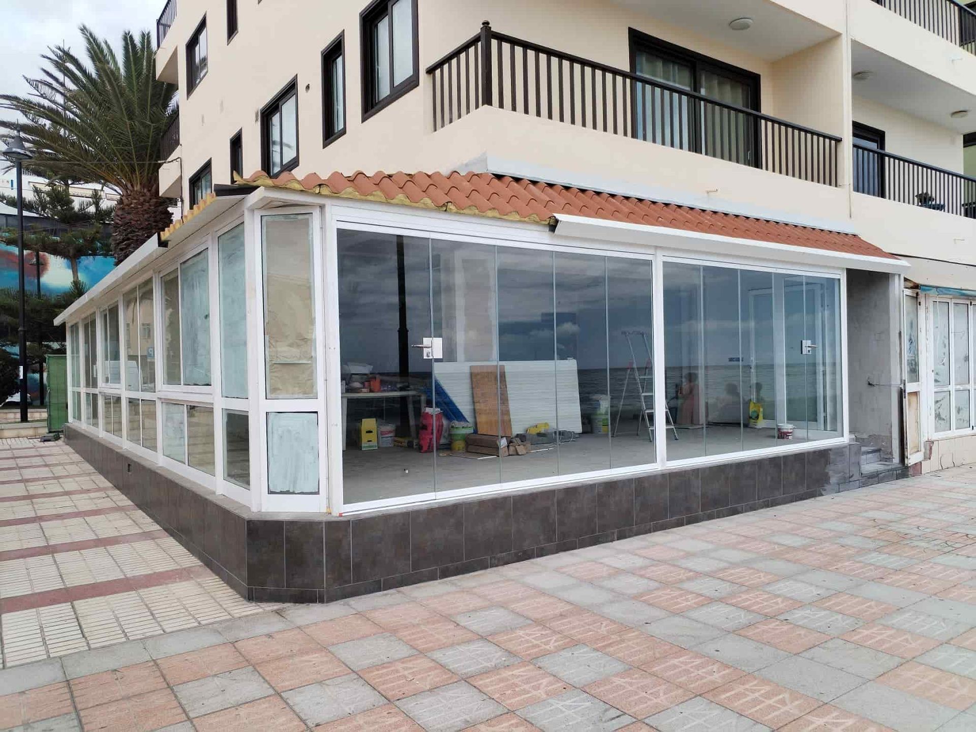 Recinto con paredes de vidrio, marcos blancos y techo de tejas rojas, situado frente a un edificio de varios pisos y una pasarela de baldosas.