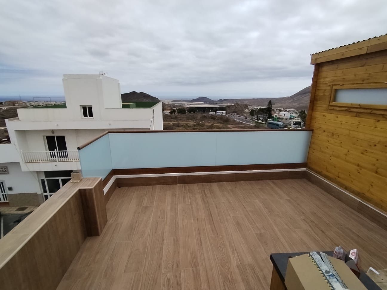 Patio en la azotea con azulejos de apariencia madera, pared azul claro y vista de una ciudad bajo un cielo nublado.